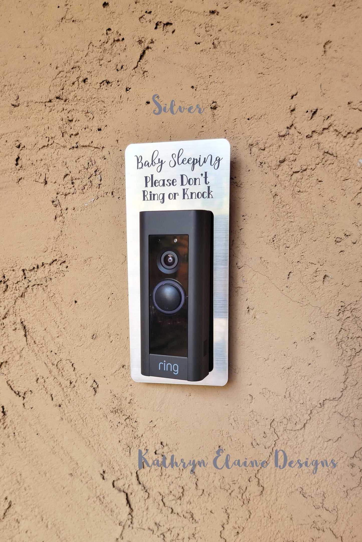 Silver laminate rectangle around Ring doorbell that says Baby Sleeping Please Don't Ring or Knock in black lettering against tan stucco background