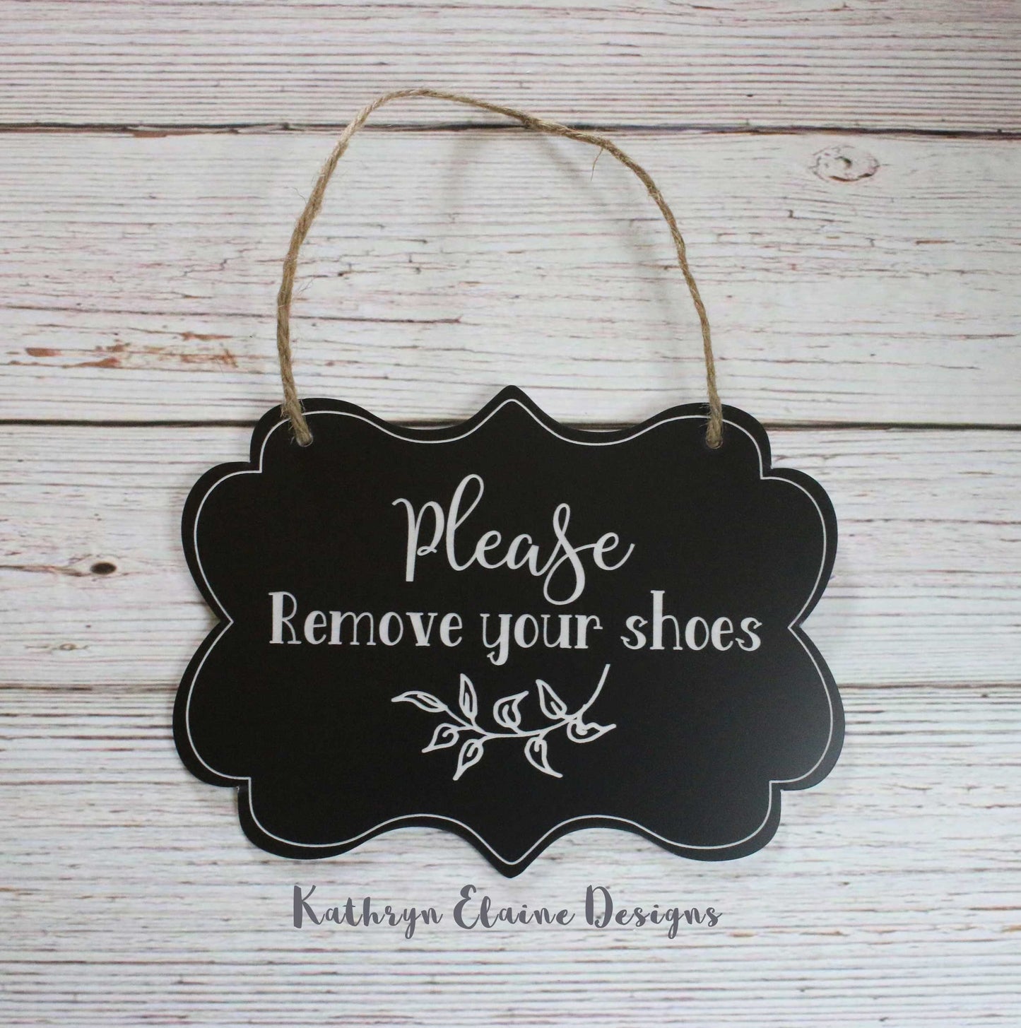 Black laminate scalloped edge sign with white lettering stating Please remove your shoes with leaf design and border on white wood background