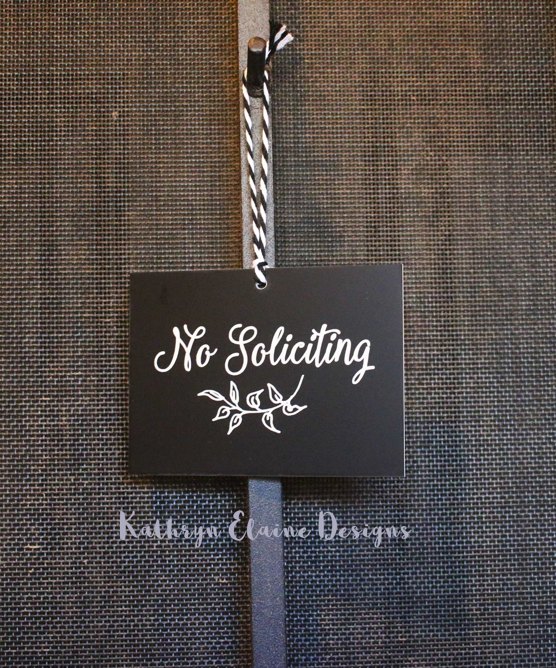 Rectangle black laminate sign with white lettering stating No Soliciting and leaf design hanging on screen door