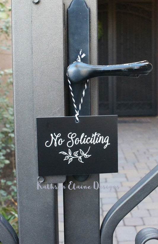 Rectangle black laminate sign with white lettering stating No Soliciting and leaf design hanging on gate.