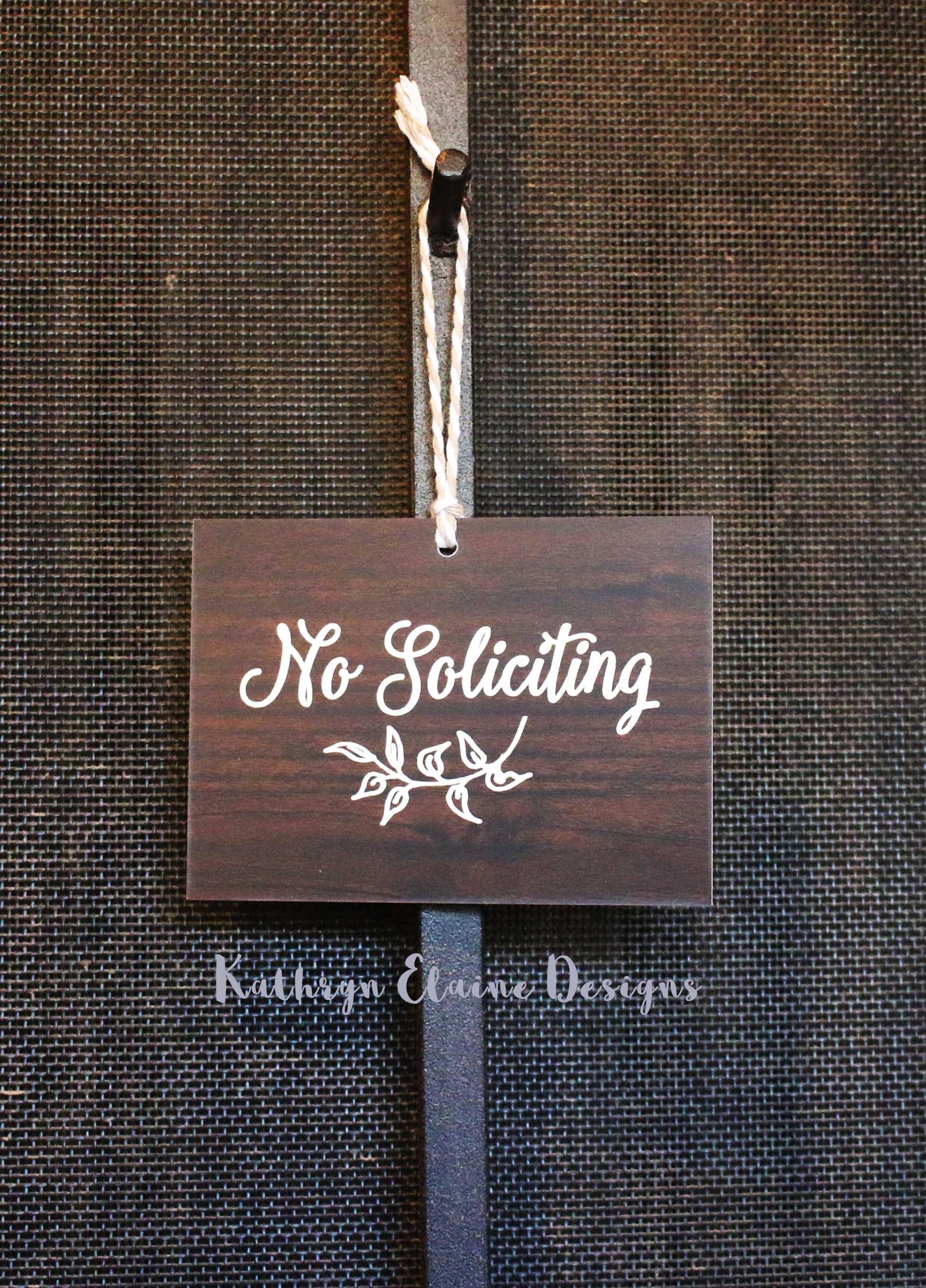 Rectangle wood grain laminate sign with white lettering stating No Soliciting and leaf design hanging on screen door