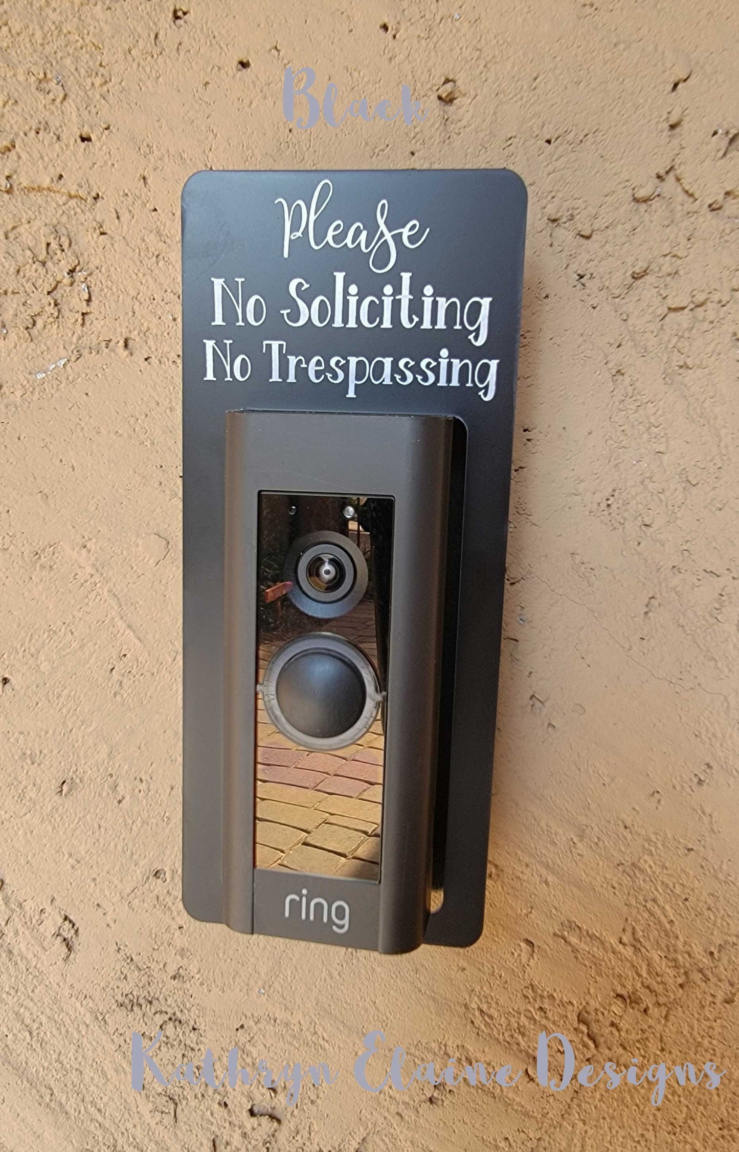 Black laminate rectangle that fits around a Ring doorbell with white laser engraved lettering stating Please No Soliciting No Trespassing on a tan background
