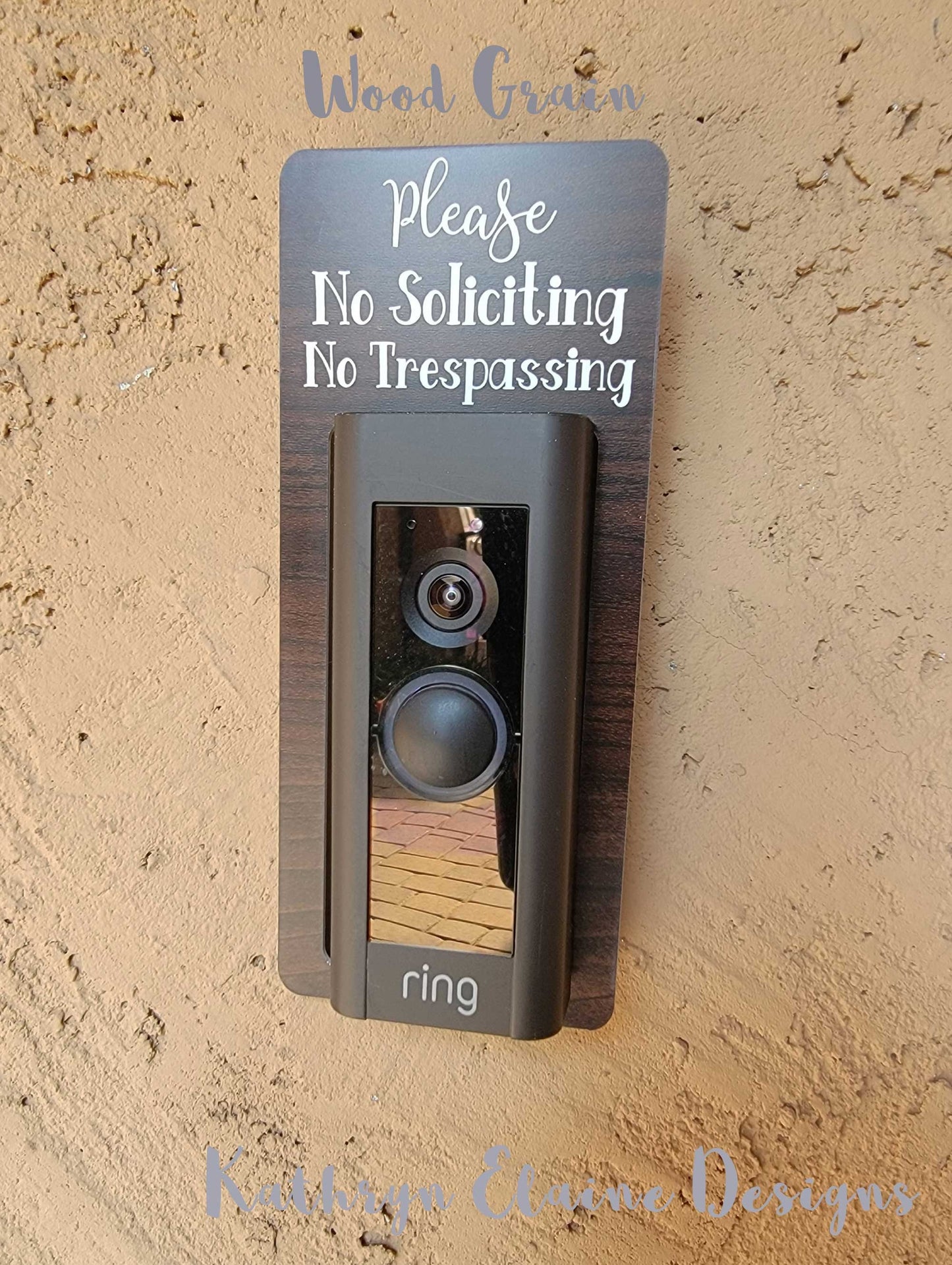Wood grain laminate rectangle that fits around a Ring doorbell with white laser engraved lettering stating Please No Soliciting No Trespassing on a tan background