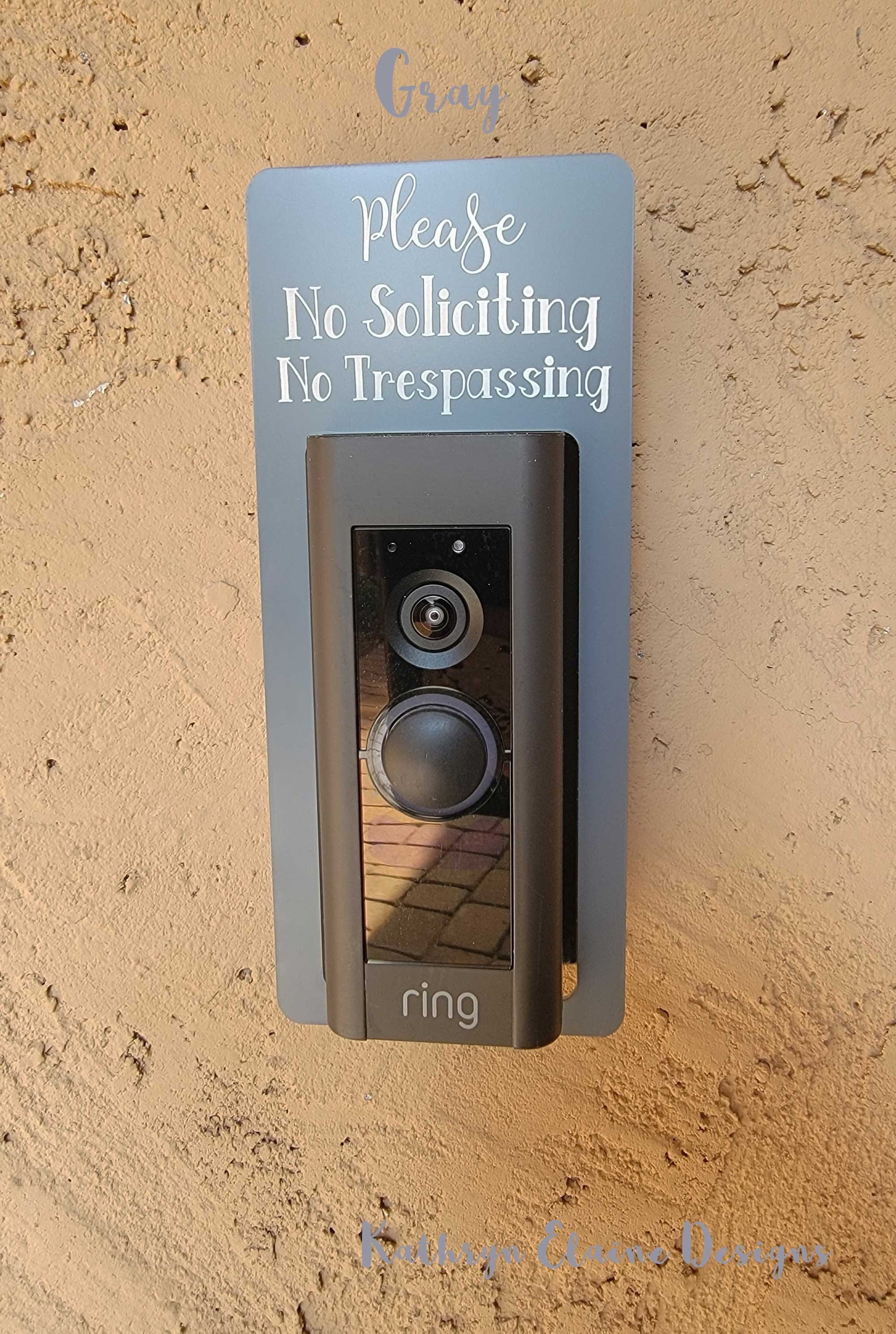 Gray laminate rectangle that fits around a Ring doorbell with white laser engraved lettering stating Please No Soliciting No Trespassing on a tan background