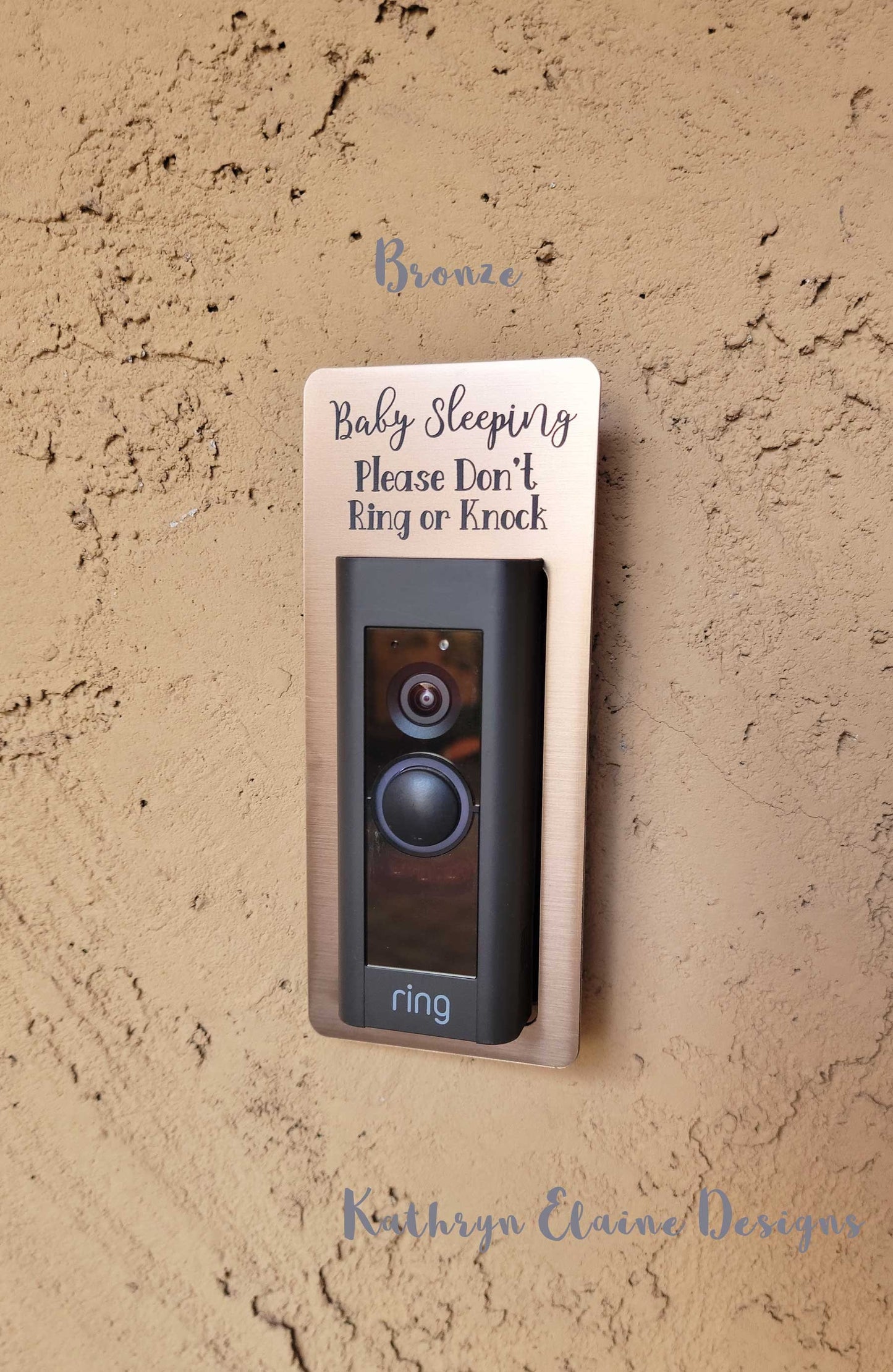 Bronze laminate rectangle around Ring doorbell that says Baby Sleeping Please Don't Ring or Knock in gray lettering against tan stucco background