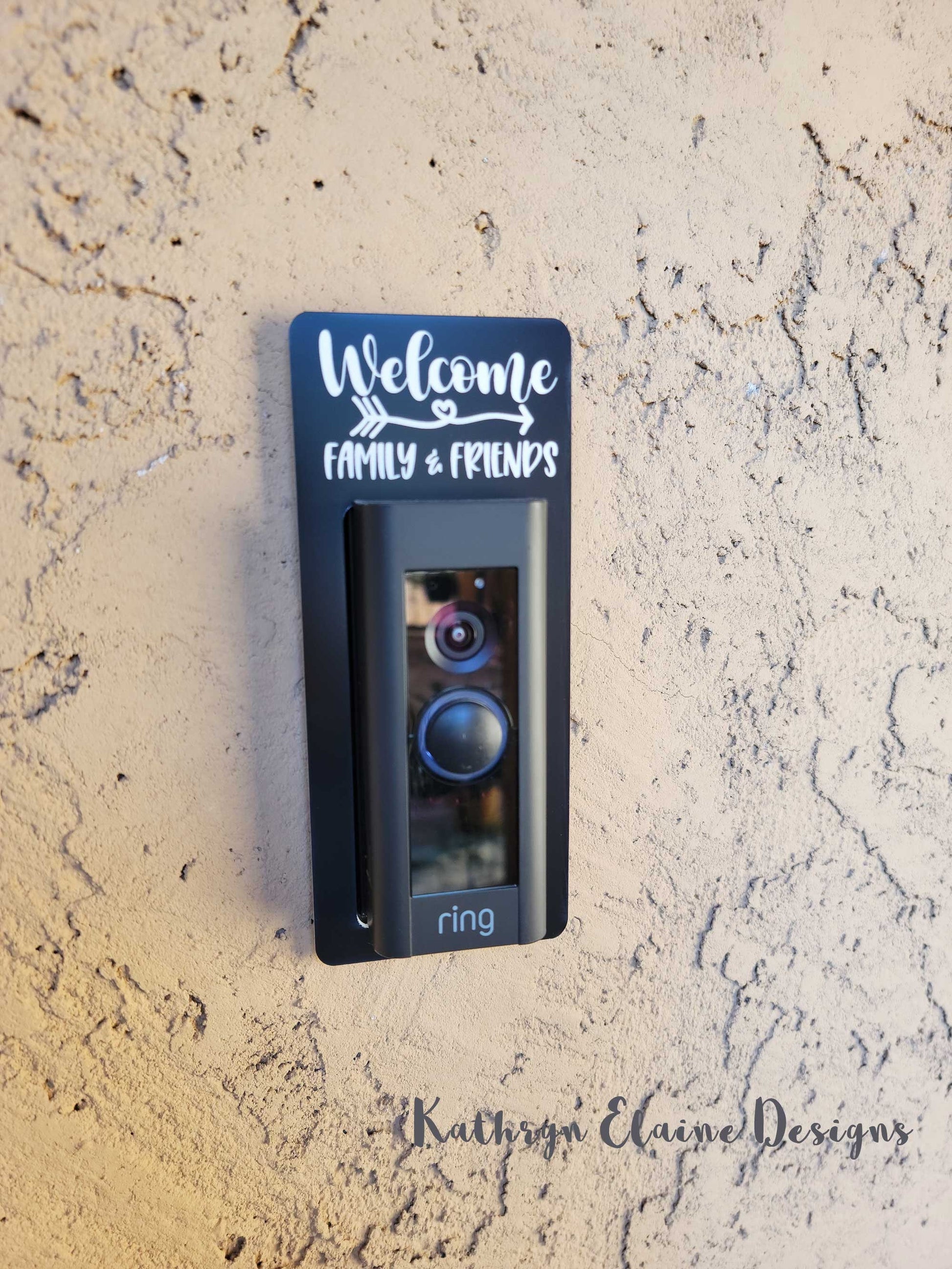 Black laminate doorbell surround with white lettering stating Welcome arrow design family and friends around Ring doorbell on tan background