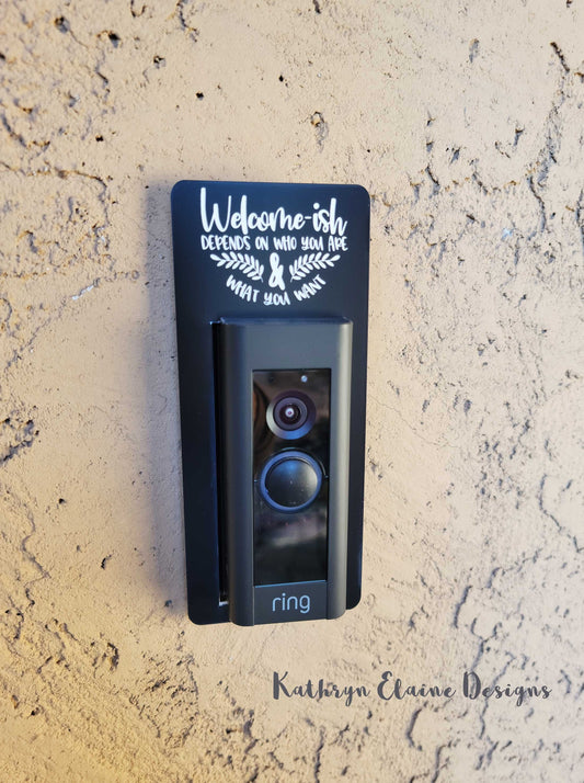 Black laminate doorbell surround with white lettering stating Welcome-ish Depends on who you are and what you want with 2 leaf designs around Ring doorbell on tan background