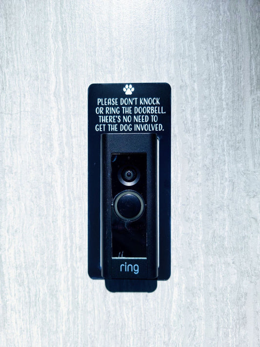 Black laminate doorbell surround with white letting stating please don't know or ring the doorbell. There's no need to get the dog involved with paw print around ring doorbell on gray background.