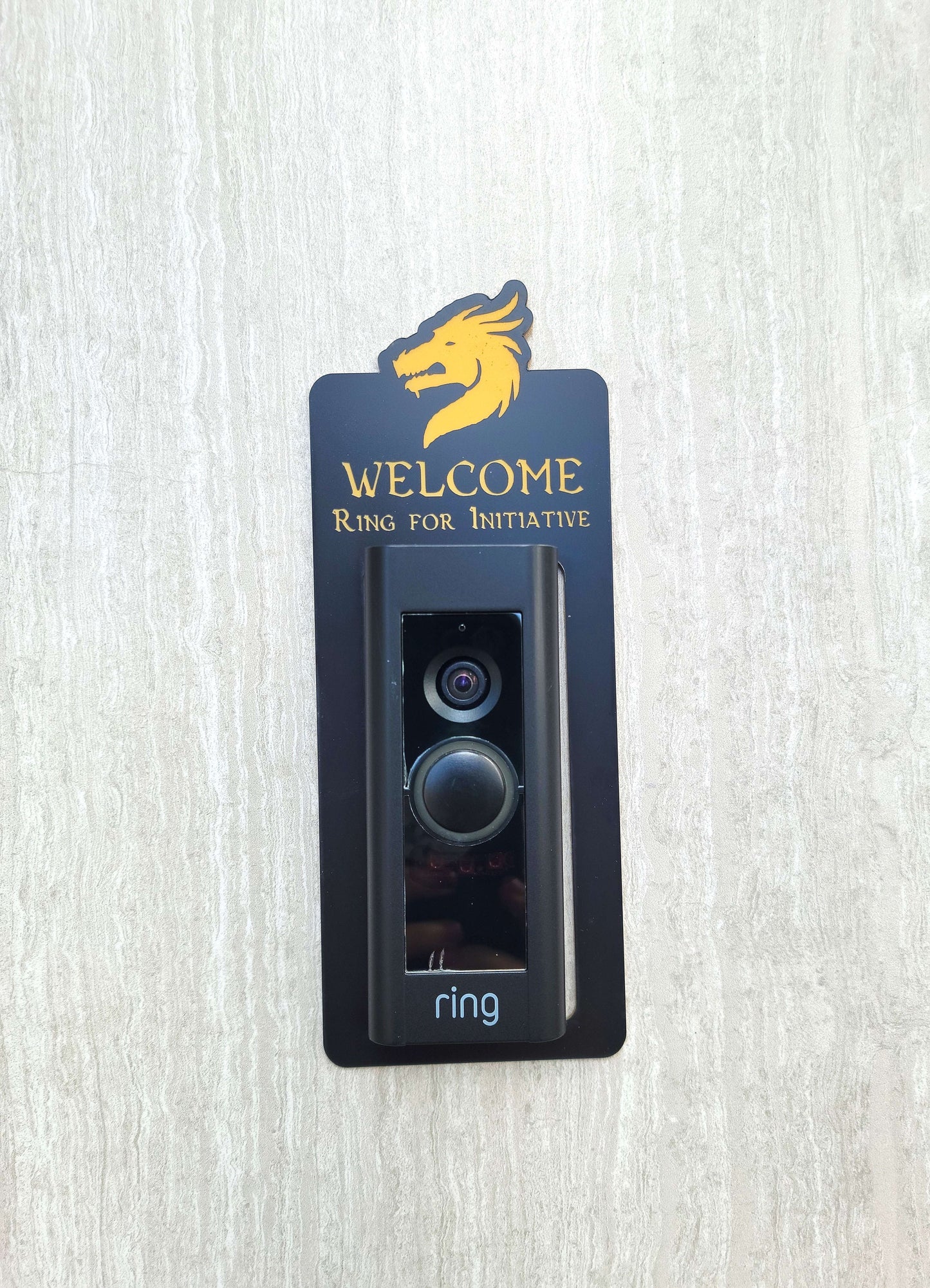 Black doorbell surround with gold dragon head and lettering stating Welcome Ring for Initiative around Ring doorbell on gray background