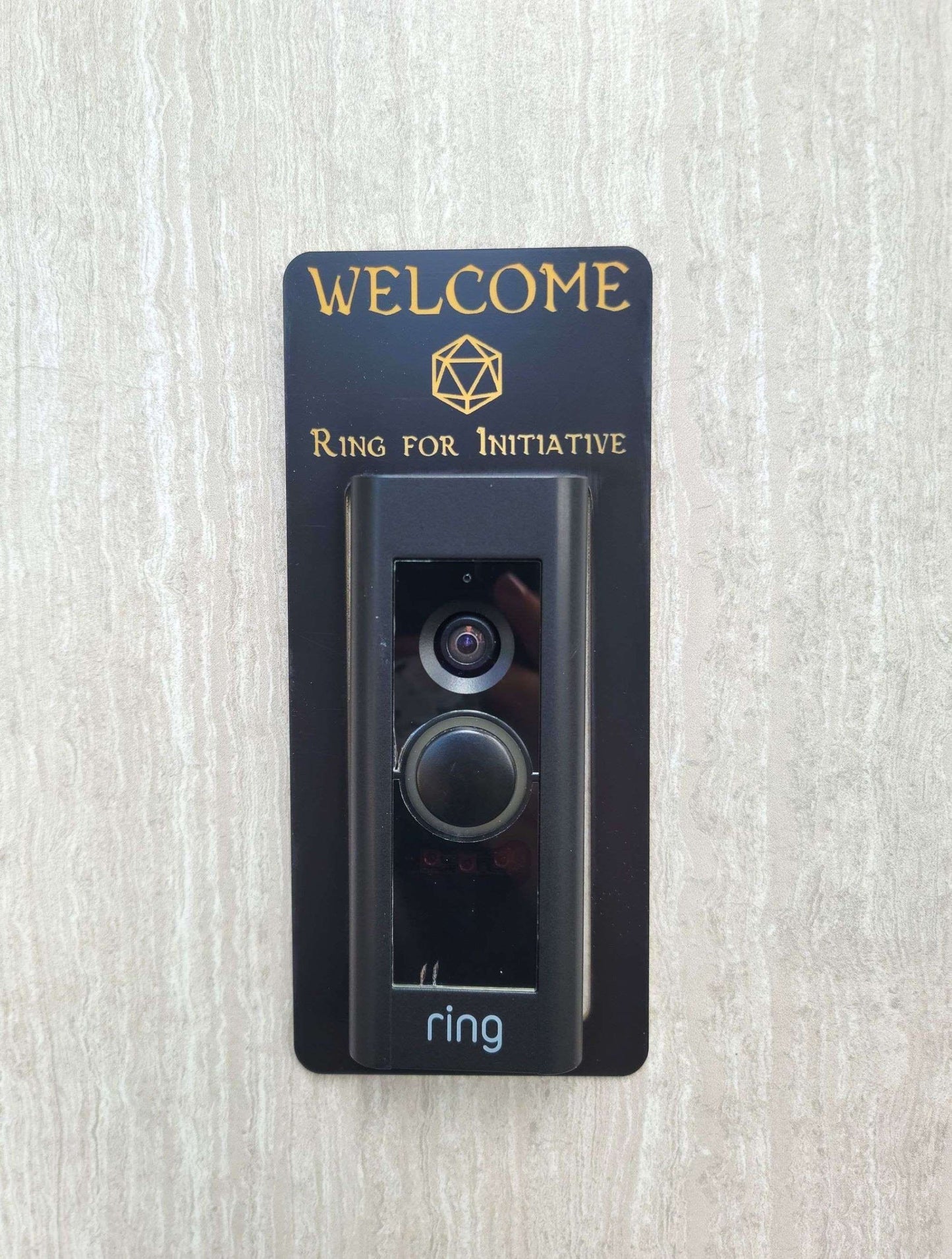 Black laminate doorbell surround with golden 20 sided dice and lettering stating Welcome Ring for Initiative around Ring doorbell on gray background