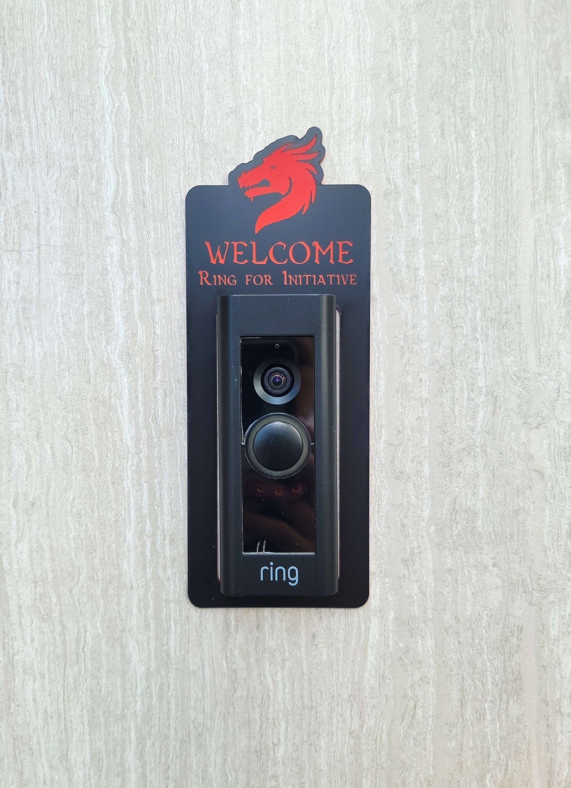 Black doorbell surround with red dragon head and lettering stating Welcome Ring for Initiative around Ring doorbell on gray background