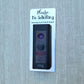 Silver laminate rectangle that fits around a Ring doorbell with black laser engraved lettering stating Please No Soliciting Seriously Don't Make It Weird on a neutral background
