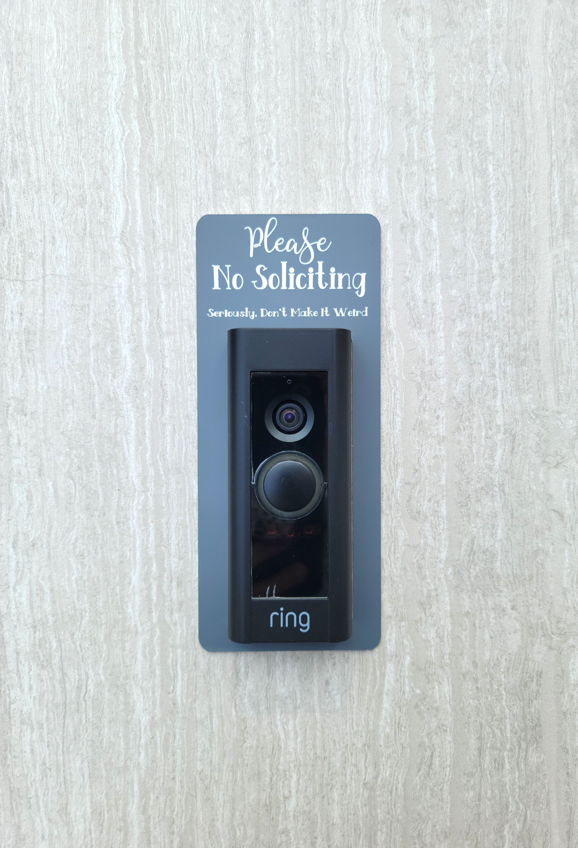 Gray laminate rectangle that fits around a Ring doorbell with white laser engraved lettering stating Please No Soliciting Seriously Don't Make It Weird on a neutral background